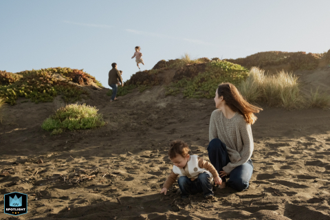 In San Francisco, California, a mom sits with her toddler on the beach, watching as her daughter joyfully jumps off the dunes into dad's arms, capturing a moment of family fun.