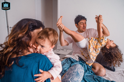 In São Paulo, Brazil, a mom cuddles a toddler, while in the background, dad plays with a young girl on the bed, capturing the joy and energy of family fun at home.