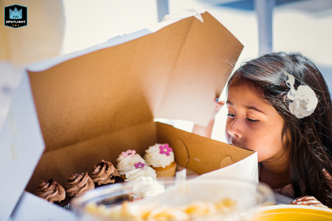 In San Francisco, California, a close-up captures a young girl lifting the cardboard lid on a box of cupcakes, her expression filled with anticipation.