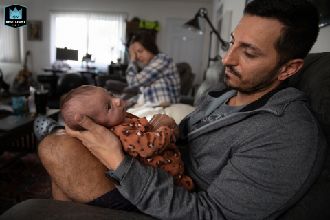 In Ventura, California, an exhausted mom yawns in the background at home while dad gently cradles their new baby, capturing the realities of early parenthood.