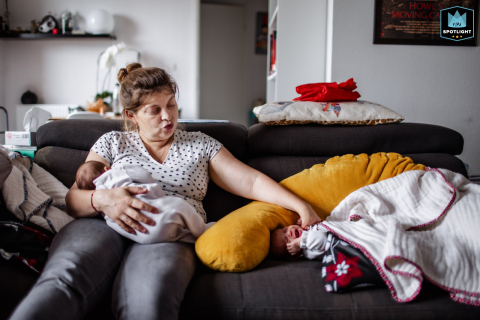 In Houston, Texas, mom multitasks with love, gently feeding one twin while soothing the other, embodying patience and care during everyday family moments.