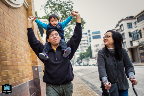 A candid San Francisco family moment: dad carries his toddler son on his shoulders along the sidewalk, while mom walks beside them pushing a stroller.