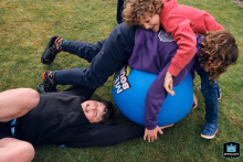 In North Yorkshire, England, children revel in rough-and-tumble fun, energetically playing with a bouncy ball on the grass, laughter echoing through their joyous outdoor game.