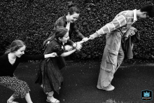In North Yorkshire, England, a black-and-white shot captures children lined up, playfully tugging an umbrella from the hand of a walking man, full of lively mischief.