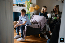 In North Yorkshire, England, a teenage boy sits alone in a chair at a party, looking bored, while women behind him chat and pour drinks together.