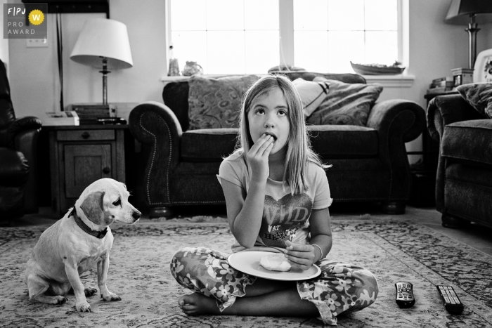 Photo by: <a href="https://www.fpja.com/documentary-family-photography-contests/julie-ambos/key-west/2034491">Julie Ambos - United States</a> A young girl enjoys a snack on the floor during a movie while her dog patiently waits for a bite in this photo captured by a Key West family photojournalist