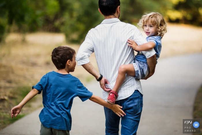 San Francisco family photojournalist captured this playful photo of a father holding his toddler son while his older son follows behind as they take off on a walk 