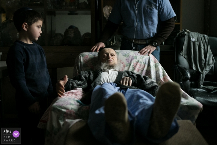 Photo by: <a href="https://www.fpja.com/documentary-family-photography-contests/polina-bulman/brooklyn/2035251">Polina Bulman - United States</a> A young boy stands next to his elderly grandfathers arm chair in this image captured by a New York City family photojournalist