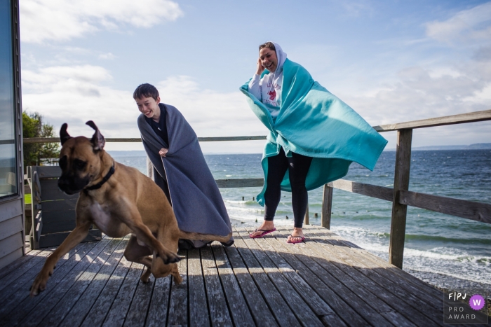 Eine Mutter und ein Sohn, die Decken tragen, wickelten um sie an einem windigen Strandtag Lachen ein, während ihr Hund über ihr Portal in dieser Fotografie läuft, die von einem Fotojournalisten der Familie San Francisco, CA hergestellt wurde.