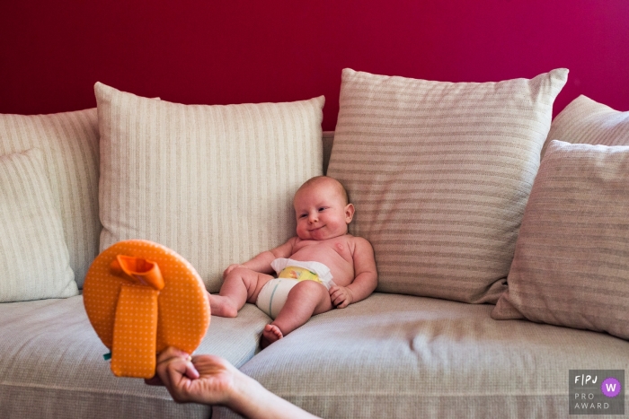 A baby sits on a couch and smiles as his mother shows him a picture in this family picture by an Antwerpen photographer. 