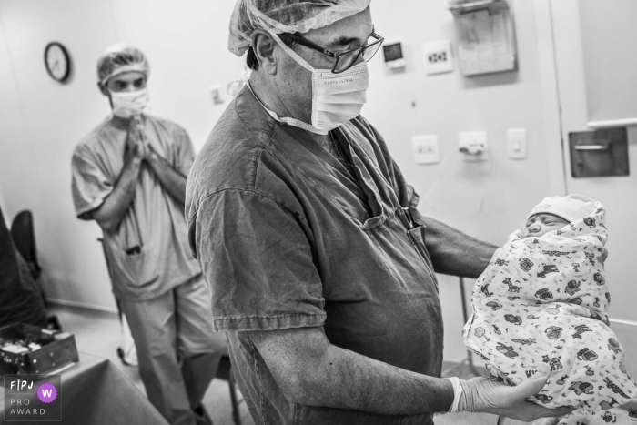 Un médecin prend un nouveau-né tandis que son père l'attend derrière lui avec enthousiasme dans cette image en noir et blanc composée par un photographe de naissance de style documentaire, Rio de Janeiro, Brésil.