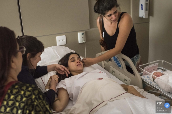 A family comforts a new mother in the hospital as her newborn sleeps next to her in this photo by a Rio de Janeiro, Brazil birth photographer.