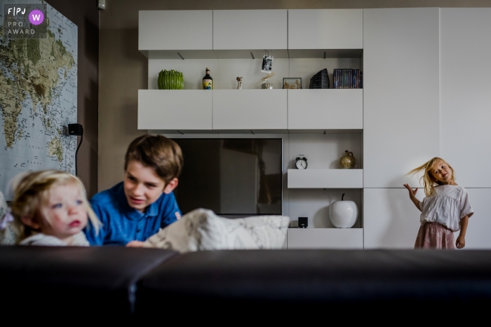 A brother and sister sit on the couch together as their other sister flips her hair in the background in this photograph by a Limburg documentary family photographer. 