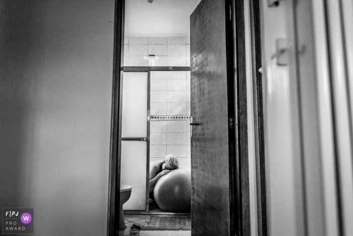 Une femme s'appuie contre une balle de yoga sous la douche lors de son accouchement à la maison, sur cette photo en noir et blanc prise par un photographe de naissance à Sao Paulo, au Brésil.