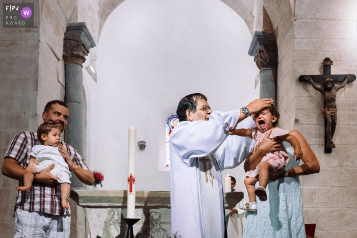 A priest attempts to bless an unhappy little girl as her mother holds her in this photograph created by a Paris, France family photojournalist. 