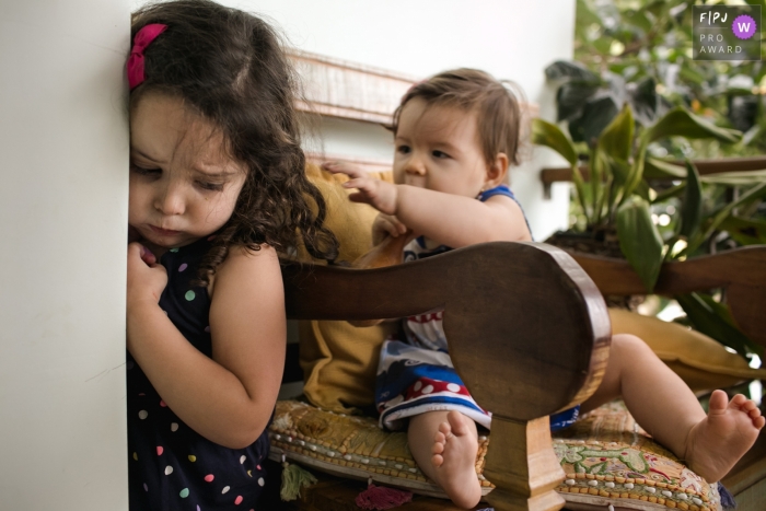 A little girl stands against a wall as her baby sister reaches for her from a chair in this FPJA award-winning picture by a Minas Gerais family photographer.