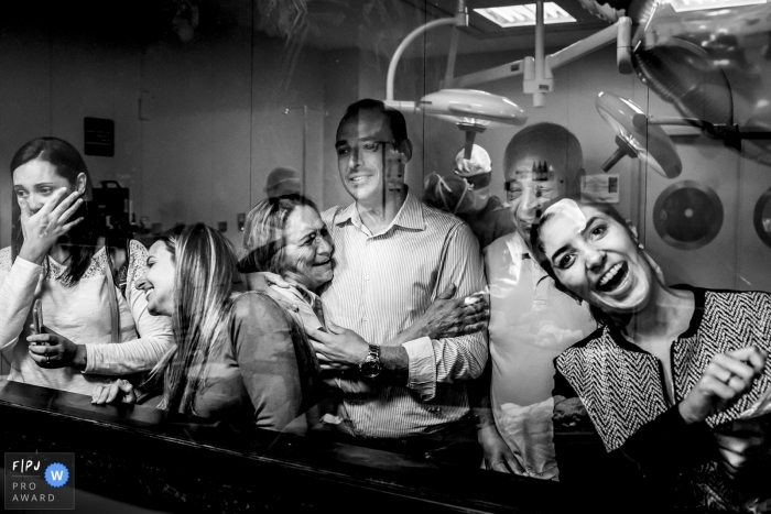 A family looks through the hospital window to see their newborn family member in this black and white photo by a Minas Gerais, Brazil birth photographer.