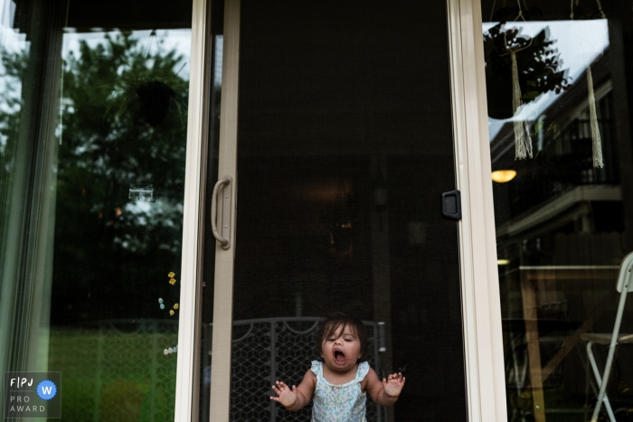 Une petite fille fait des grimaces sur une porte moustiquaire dans cette photo récompensée par la photo par un photographe de famille documentaire du Overland Park, KS.