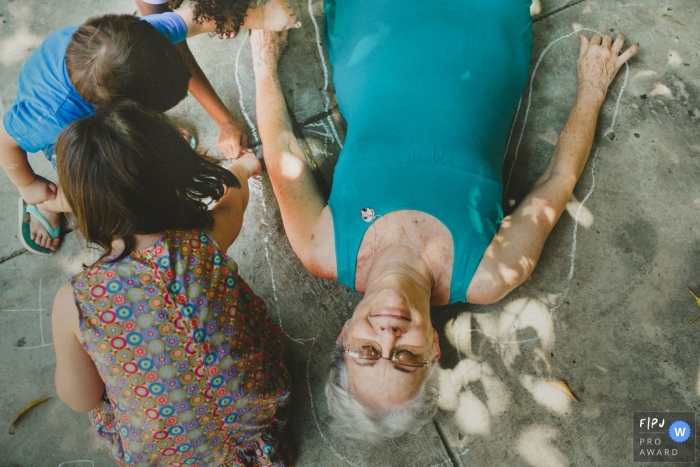 Photo by: <a href="https://www.fpja.com/documentary-family-photography-contests/aline-lelles/rio-de-janeiro/2063211">Aline Lelles - Brazil</a> Three children outline their grandmother in chalk on the driveway in this photo by a Rio de Janeiro, Brazil award-winning family photographer.