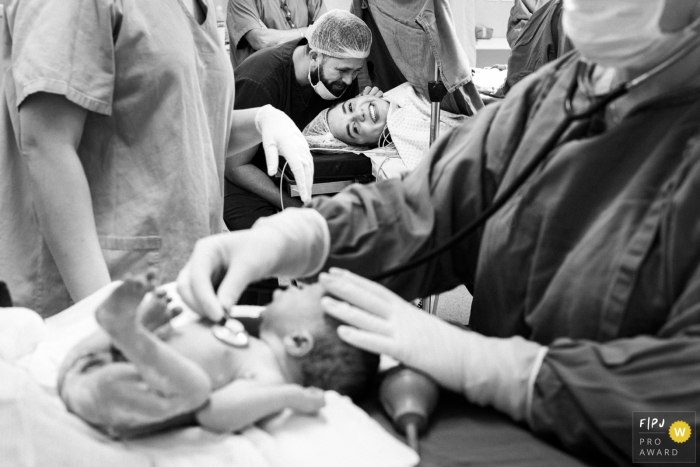 A mother lays on a hospital bed looking over at her newborn as the doctor checks her heartbeat in this black and white photo by a Minas Gerais, Brazil birth photographer.