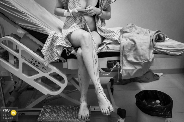 A pregnant woman sits on the edge of a hospital bed as she checks her phone in this black and white birth photo captured by a Minas Gerais, Brazil documentary photographer.