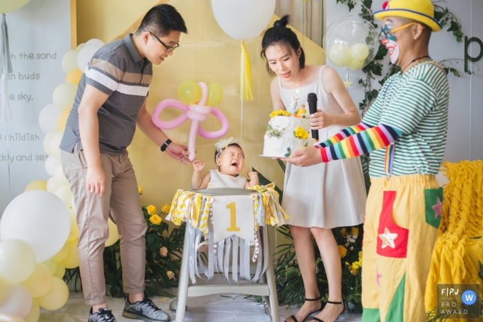 A baby girl cries at her first birthday when she sees a clown carrying the cake in this photo recorded by a Zhejiang, China award-winning, documentary-style family photographer. 