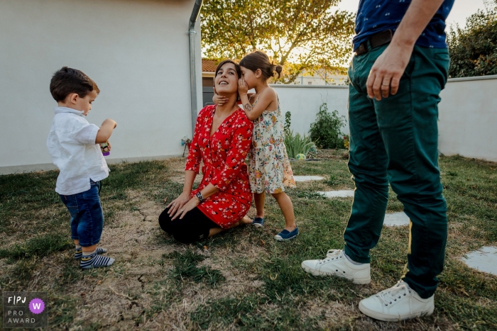 A girl whispers into her mother's ear as the family spends time outside in this Family Photojournalist Association contest awarded photo created by a Haute-Garonne family photographer. 