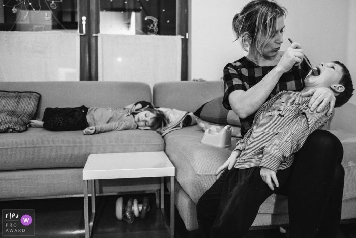 A mother brushes her son's teeth as her daughter sleeps on the couch in this photo recorded by an Antwerpen award-winning, documentary-style family photographer. 