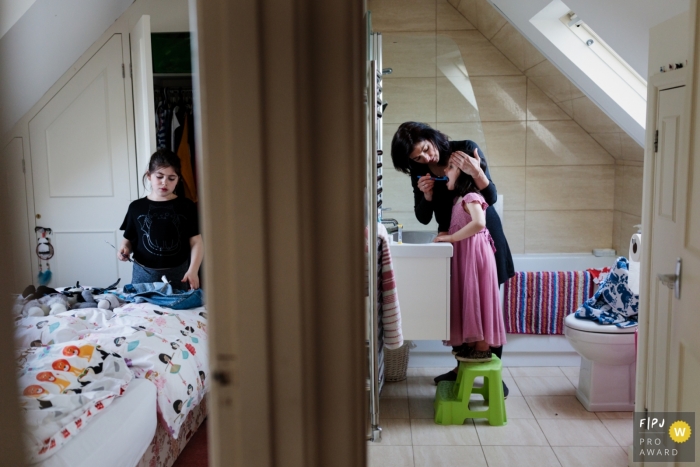 A mother helps her daughter brush her teeth in the bathroom while her other daughter cleans her room in this documentary-style family photo captured by a Kent, England photographer. 