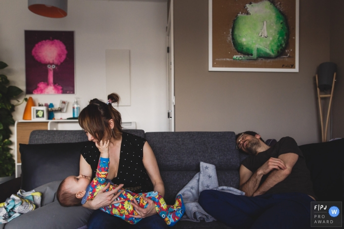 Une mère joue avec son fils tandis que son mari dort à côté d'eux sur le canapé dans cette image familiale de style documentaire enregistrée par un photographe de Paris, France.