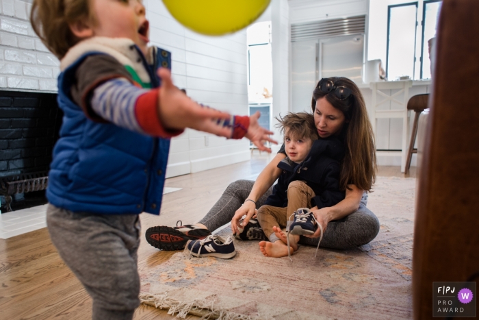 Photo by: <a href="https://www.fpja.com/documentary-family-photography-contests/melissa-mcclure/los-angeles/2068096">Melissa McClure - United States</a> A mother helps her son put his shoes on as his brother plays with a ball in this photograph by a Los Angeles, CA documentary family photographer.