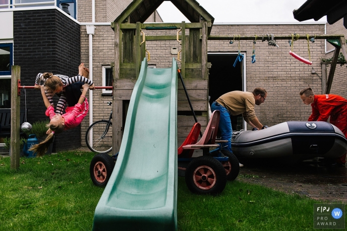 Photo by: <a href="https://www.fpja.com/documentary-family-photography-contests/liesbeth-parlevliet/zuid-holland/2068416">Liesbeth Parlevliet - Netherlands</a> Two girls play on a swing set as their father and brother move an inflatable boat in this FPJA award-winning picture by a Netherlands family photographer.