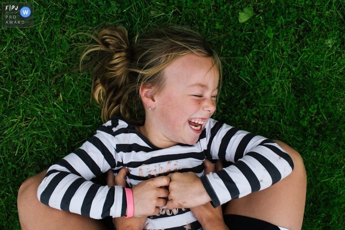 A little girl lays in the grass laughing as someone tickles her in this documentary-style family photo captured by a Netherlands photographer. 