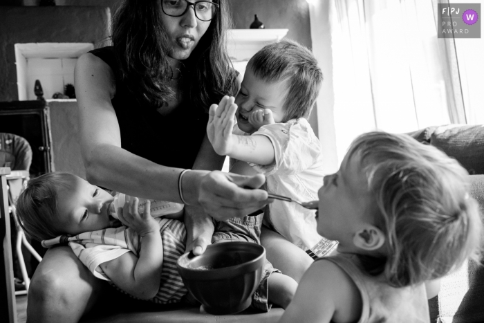 A mother tries to feed her three young children simultaneously in this photograph by a Nantes documentary family photographer. 