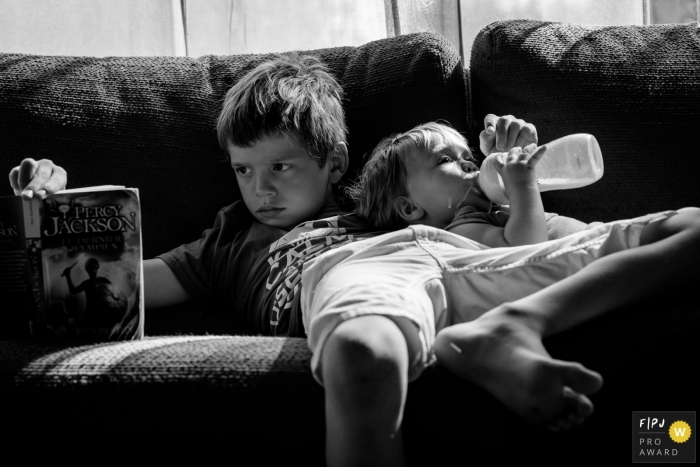 A boy reads on the couch while his little brother drinks his bottle next to him in this FPJA award-winning image captured by a Nantes family photographer. 