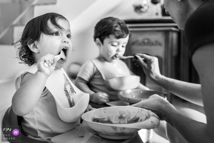 Photo by: <a href="https://www.fpja.com/documentary-family-photography-contests/priscilla-mondo/rio-de-janeiro/2068836">Priscilla Mondo - Brazil</a> A little girl eats her lunch while her mother feeds her brother in this documentary-style family photo captured by a Sao Paulo, Brazil photographer.