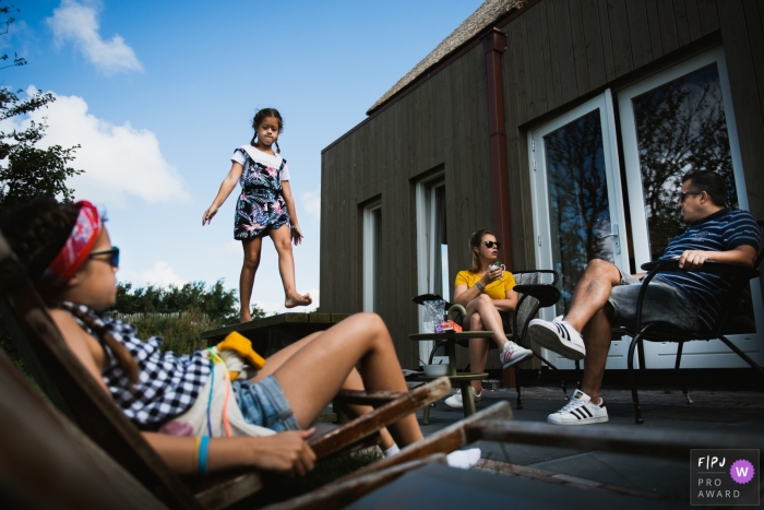 Ein Mädchen geht auf einen Tisch, während ihre Familie draußen in dieser Family Photojournalist Association sitzt, die von einem Amsterdamer Dokumentarfamilienfotografen mit einem Foto ausgezeichnet wurde.