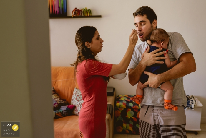 A woman feeds her husband as he uses both arms to hold their baby in this photo recorded by a Rio de Janeiro, Brazil award-winning, documentary-style family photographer. 