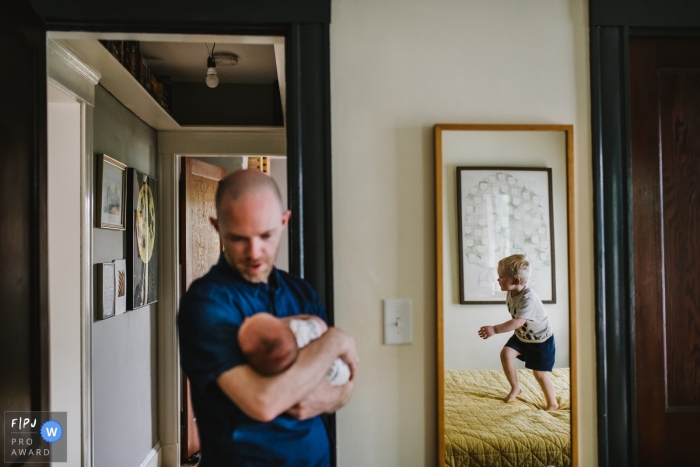 Photo by: <a href="https://www.fpja.com/documentary-family-photography-contests/korri-crowley/boston/2069151">Korri Crowley - United States</a> A father holds his baby as his son can be seen jumping on the bed in a mirror in this documentary-style family photo captured by a Boston, MA photographer.
