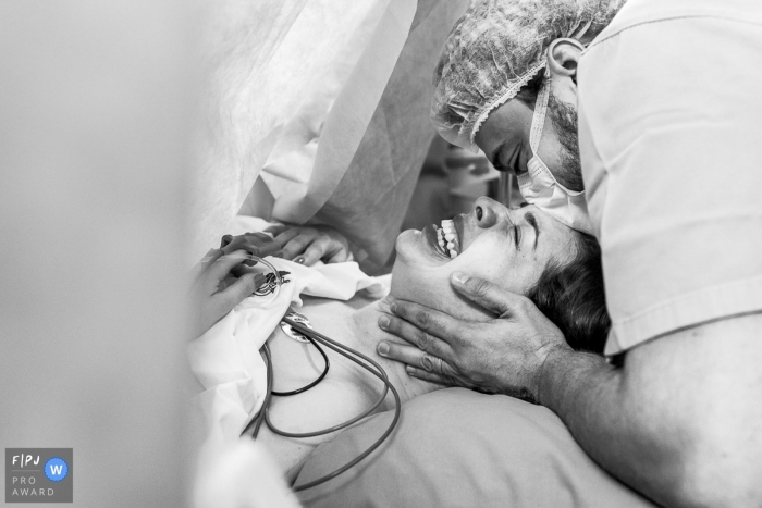 A man kisses his wife's forehead through a surgical mask after she gives birth in the hospital in this black and white image composed by a Rio de Janeiro, Brazil birth photographer. 