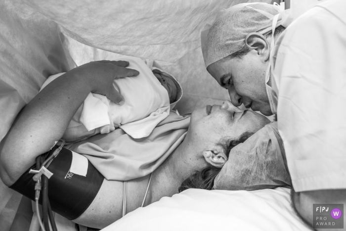 Photo by: <a href="https://www.fpja.com/documentary-family-photography-contests/claudia-ruiz/rio-de-janeiro/2069981">Claudia Ruiz - Brazil</a> A husband touches noses with his wife as she holds their newborn in the hospital in this birth photograph by an award-winning Rio de Janeiro, Brazil photographer.