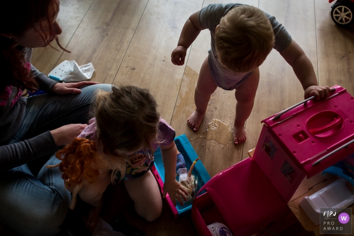 A little boy has an accident on the floor as his mother and sister play with dolls next to him in this FPJA award-winning picture by a Hollands family photographer.