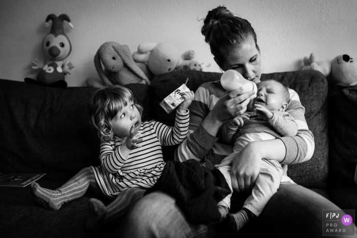 A mother tries to bottle feed her baby as her young daughter holds up her juice box in this documentary-style family photo captured by a Netherlands photographer. 