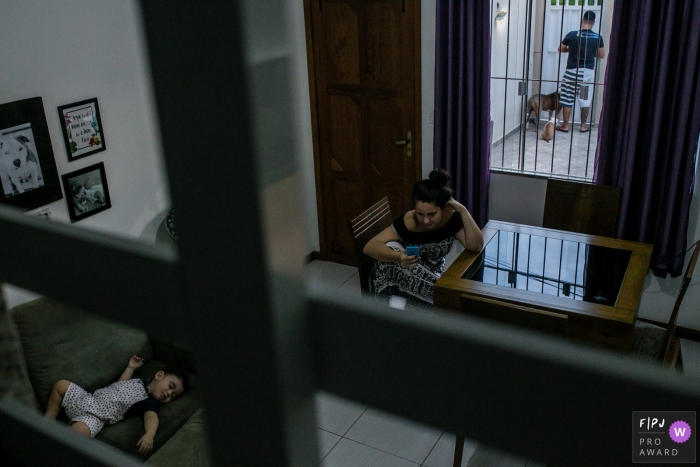 Photo by: <a href="https://www.fpja.com/documentary-family-photography-contests/raphael-bozeo/maca%C3%A9/2071076">Raphael Bozeo - Brazil</a> A mother sits at a table while her son sleeps on the couch in this photograph by a Rio de Janeiro documentary family photographer.