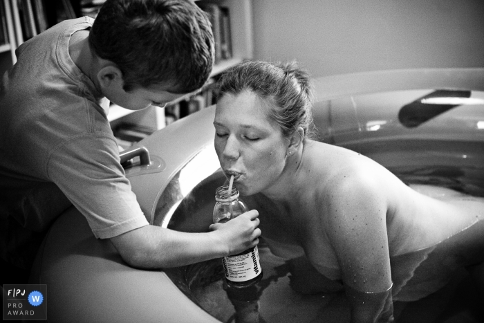 A boy gives his mother a drink during her at-home water birth in this black and white photo by a documentary-style Fort Wayne, IN birth photographer.