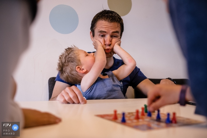 Photo by: <a href="https://www.fpja.com/documentary-family-photography-contests/huibert-van-rossum/zuid-holland/2071991">Huibert van Rossum - Netherlands</a> A young boy squeezes his father's cheeks as people sit at a table playing a game in this photo by a Netherlands family photojournalist.