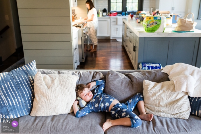 Photo by: <a href="https://www.fpja.com/documentary-family-photography-contests/david-clumpner/missoula/2072471">David Clumpner - United States</a> A boy lays sprawled on the couch as his mother cooks in the kitchen in this photo by a Missoula, MT family photojournalist.