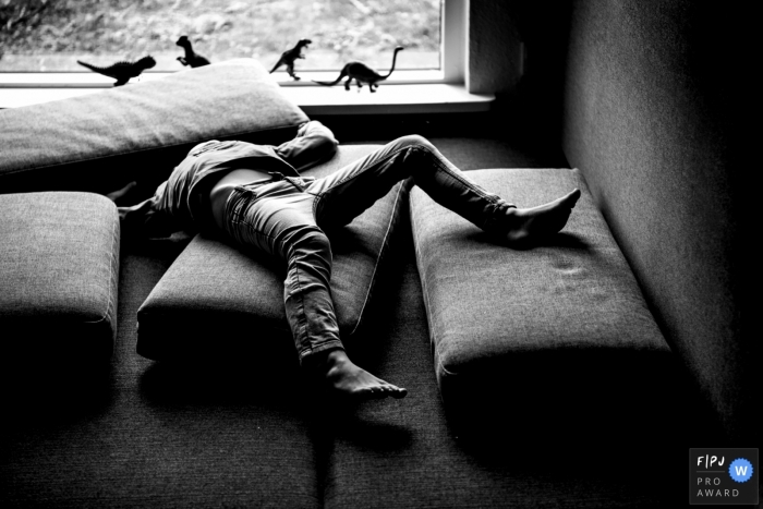 A child plays with cushions as toy dinosaurs stand lined up on the window sill in this black and white photo by a Copenhagen, Denmark family photojournalist.