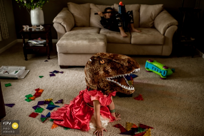 A little girl in a princess dress wears a T-Rex head as her brother sits on the couch in this photo by a Spokane, WA family photojournalist. 