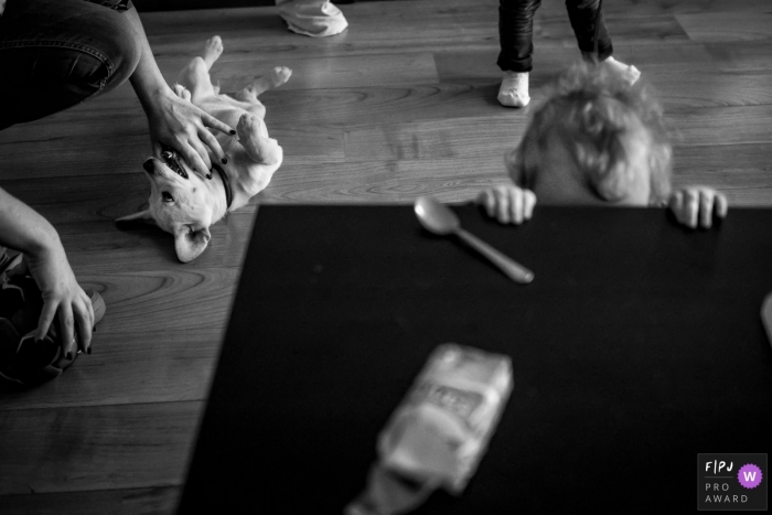 Photo by: <a href="https://www.fpja.com/documentary-family-photography-contests/katrin-k%C3%BCllenberg/dusseldorf/2073756">Katrin Küllenberg - Germany</a> A mother pets a dog in the background as her daughter attempts to peek over a table in this black and white photo by a Dusseldorf family photojournalist.